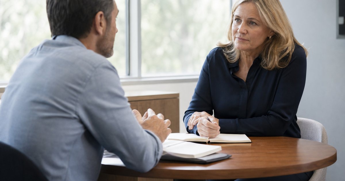 Two people in a quiet office having a serious one-on-one conversation at a small round table, notebooks open, a window with soft daylight behind them